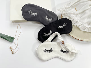 Three luxury silk sleep masks in black, dark gray, and white, each embroidered with decorative eyelash designs, displayed on a light background.