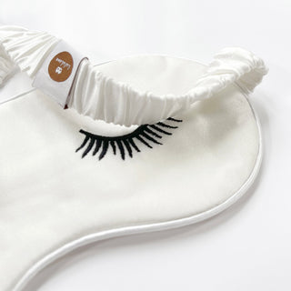 Luxurious white silk sleep mask with elegant black embroidered eyelash design and elastic band featuring a small branded tag