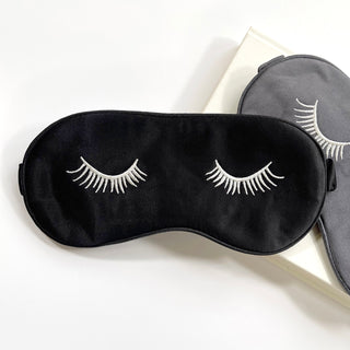 Luxury black silk sleep mask with white embroidered eyelashes design, displayed with its gray reverse side on a cream background