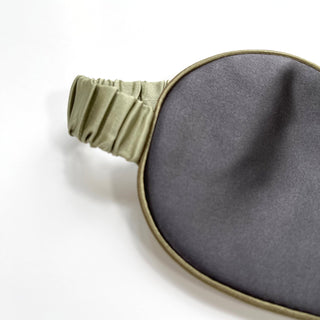 Luxurious double-sided sleep mask featuring deep charcoal silk and soft sage green elastic band with elegant piping detail