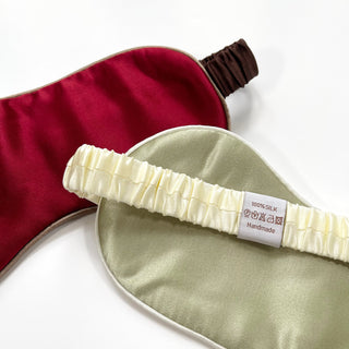 Two luxury silk sleep masks - one deep red with brown elastic band and one sage green with cream elastic - showing 100% silk handmade tag