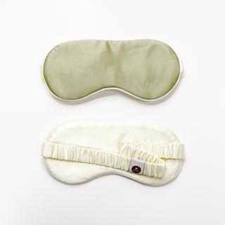 Double-sided luxury silk sleep mask showing sage green side on top and cream side below with elastic band and brand tag on white background.