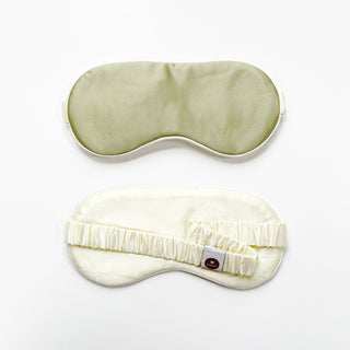 Double-sided luxury silk sleep mask shown in sage green and cream colors, featuring an elastic band and brand tag for comfortable nighttime wear