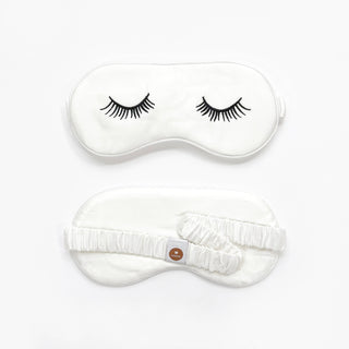 Double-sided white silk sleep mask with embroidered eyelash design on front and elastic band with brand tag on back, displayed on white background
